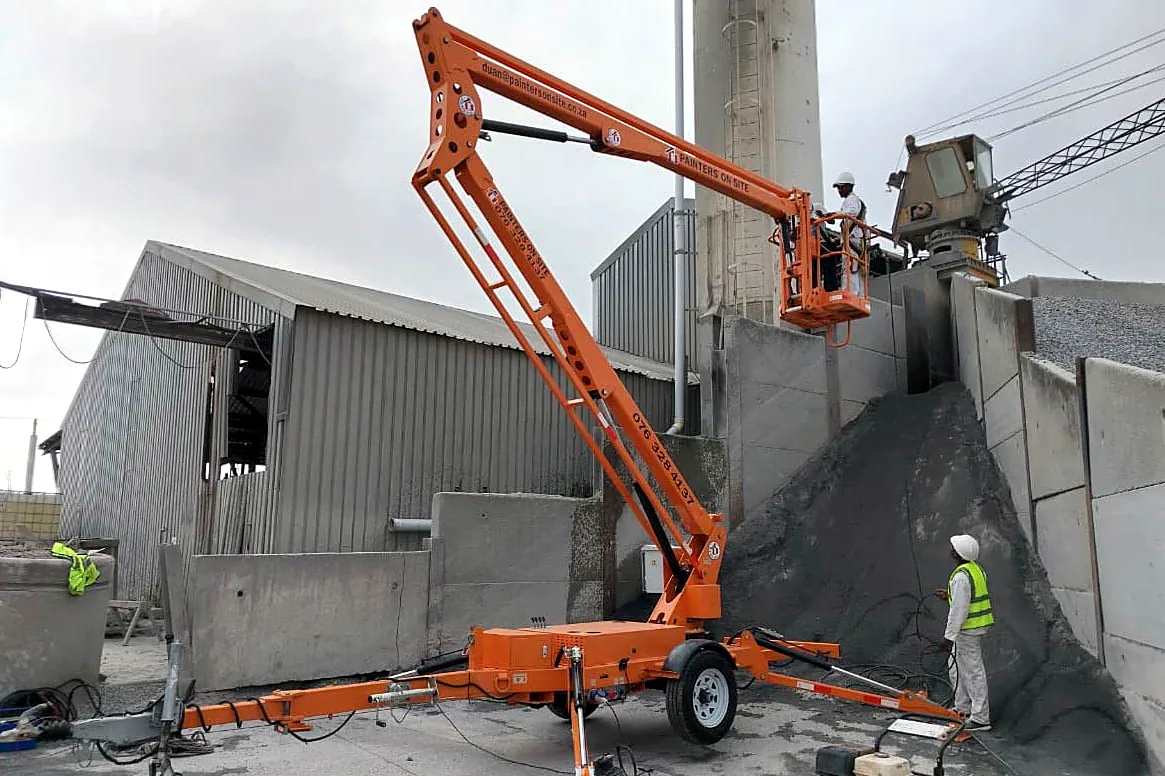 MCM towable cherry picker raised beside a cement plant silo with ground crew in high-visibility vests