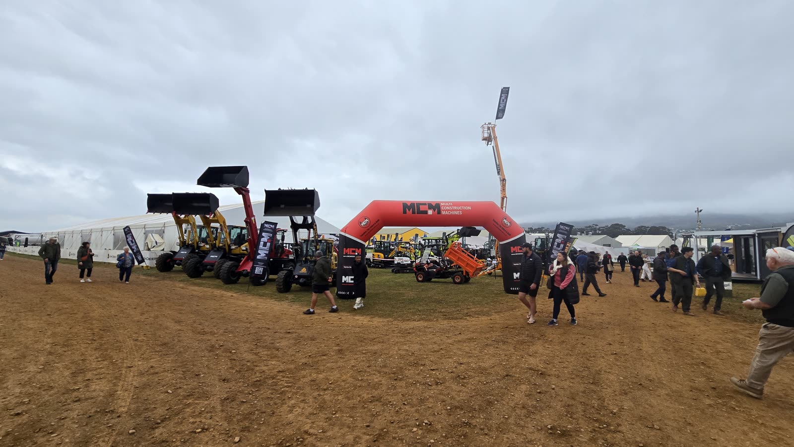 Visitors walking past the MCM entrance arch at NAMPO Cape 2026, with front end loaders, the T250 telescopic loader, Aerobs UTV and TCP16 cherry picker on display
